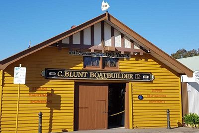 Colour photo of the exterior of a weatherboard shed painted mustard yellow with a peaked roof. Signage on the front above the entry with the words C Blunt Boatbuilder.