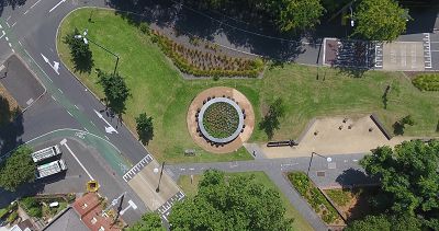 An aerial photograph of a 7m diameter round timber table located in a public park. The table is surrounded by lawn and in its centre is a garden.