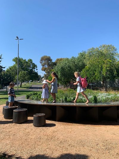 Three children walking on a round timber table set within a park. A circular herb garden is at the centre of the table.