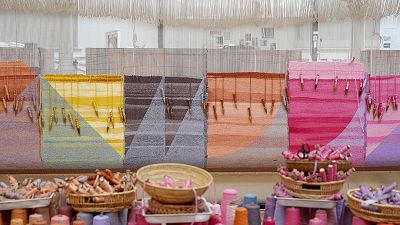 A close up of the working loom with geometric shapes - mostly triangles with colours of yellow, grey, mango and hot pink. Straw baskets in the foreground with wool and bobbins.