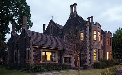 A view of The Gate House Lodge at Melbourne General Cemetery from inside the grounds. It is dusk and the windows are lit up inside