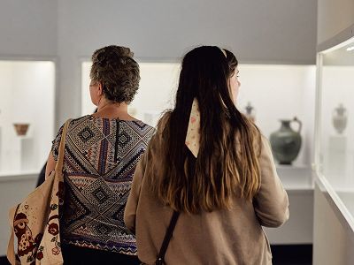 The back of an older woman in a patterned shirt and a young woman in a knit sweater, who are both looking ahead towards Museum cabinets which are out of focus. The silhouette of a vase and a bowl are visible in the glass cabinets.