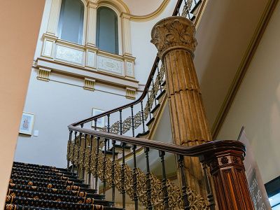 An upwards view of the ornate staircase in the foyer of the Hellenic Museum. Its dark wooden pillar and balustrade are visible in the foreground. Tall ceilings are visible in the background, with a set of large, frosted windows with ornamentation on the back wall.