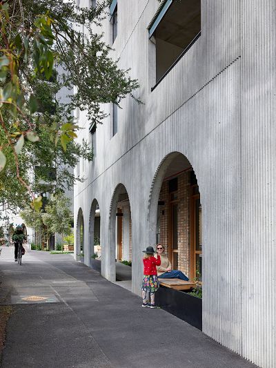 the image is dominated by the fluted concrete facade of Nightingale Anstey which features a series of archways along a pedestrian colonnade, and a blonde woman sits on a bench in the closest archway smiling at a young child in a red jumper, while a cyclist rides towards us along the cycle path to the left of the picture.