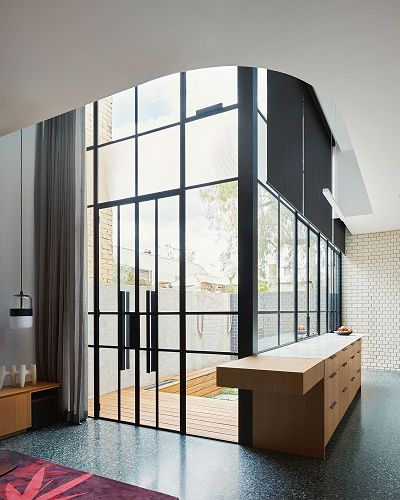Image taken from the interior of a house looks through a set of almost five-metre-high steel and glass doors and windows, towards a courtyard. To the right, there is a sleek, marble and timber standalone kitchen bench inside the kitchen, with a curved white brick wall beyond it. To the left, a light hangs over an abstract sculpture on a timber side table.