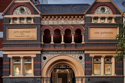 Photograph of the front of a building, dark brick in colour with cream accents. The building is symmetrical, with a central arched entrance, decorative arched balcony, and pointed roof facing the viewer on either end.