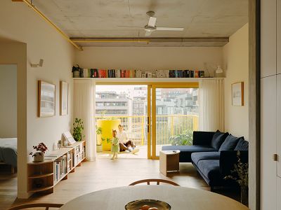 an apartment living room with a circular dining table in the foreground, a deep blue couch against the wall on the right, and a view through sliding doors out the a balcony at the back of the shot, where a parent sits on the pavers and watches their toddler who is wearing a green dress