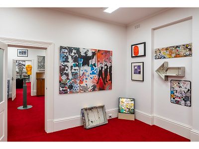 Display of street art objects as part of the City of Melbourne's Art and Heritage Collection store. A three panel spray-painted and colourful artwork hangs on the centre wall and features images of waratah flowers, bushranger Ned Kelly and a group of children. To the right and below are 7 street art related objects and artworks including a square and arrow shaped light box and some painted artworks. On the left hand side is the doorway to the next room, which features historical carpark ticket machines and a framed photograph of City of Melbourne ticket inspectors.