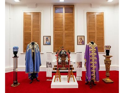 Display of objects relating to Melbourne's Moomba festival in the City of Melbourne's Art and Heritage Collection store. Carpet on floor is red and shutters behind objects are dark brown wood. At the centre is the Mikoshi shrine and is brown and red in colour with gold bird detailing. On the left is a light blue and gold Moomba Queen crown, robe and portrait of a Moomba queen. On the right side is a purple and gold Moomba King crown, robe and portrait of a Moomba King. Both crowns are displayed on high stands.
