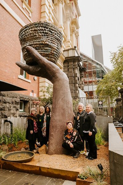 The group of artists who created the Creative Resilience sculpture stand in front and beside the sculpture smiling at the camera. They are standing in the sculpture garden forecourt located at the front of the Queen Victoria Women's Centre building.