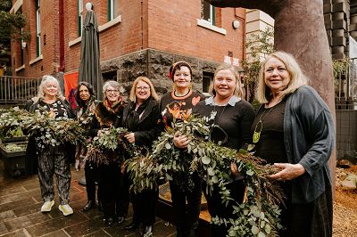 The group of artists from the Ngardang Girri Kalat Mimini (NGKM) collective are standing in a row with Natalie Hutchins, the Minister for Women holding a wreath garland of native flowers in front of the Creative Resilience sculpture.