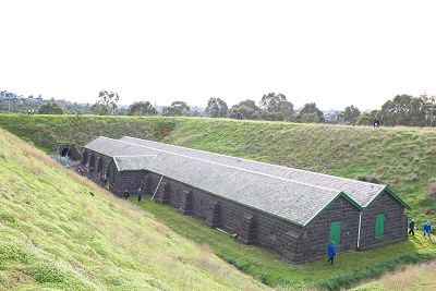 Exterior of bluestone magazine surrounded by tall grassy blast mounds. People are exploring the outside of building at ground level. People are walking around on top of the mounds.