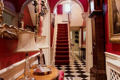 A photograph looking through a narrow hallway. The wallpaper is deep red and the floor is checkered black and white. There is a small wooden side table with lamps to the left, and a large vintage clock on the right.
