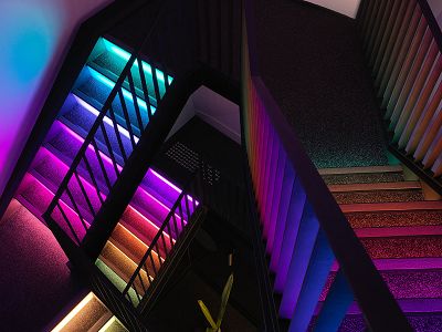 A birds-eye-view photograph of a staircase which traverses three floors. The steps are lit-up with multi-coloured lights.