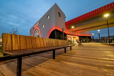 The new, modern Coburg station at dusk. The ‘Coburg’ signage and architectural design features are lit up in red. A timber deck stretches to the station forecourt with steel and timber seating beside a garden bed. The elevated rail is heading north towards Batman Station.
