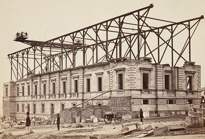 Old Treasury Building under construction, the external lower 2 levels are completed, while a metal frame juts above with a long crane suspended across the building attached to the outer edge.