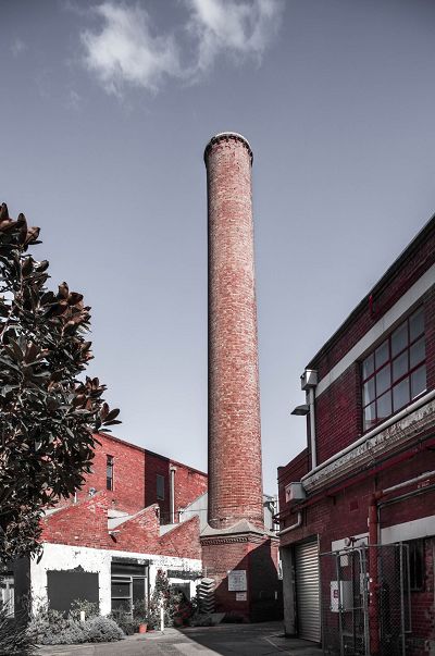 Streetscape shows a red brick chimney soaring over two red industrial brick buildings. One has a sawtooth roof.