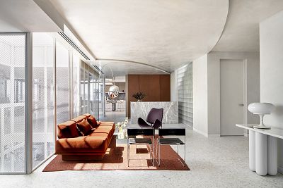Light streams through a window into a sleek, contemporary waiting room with a brown couch and a few chairs around a coffee table with flowers on it. Behind is a marble reception desk in front a dark timber wall.