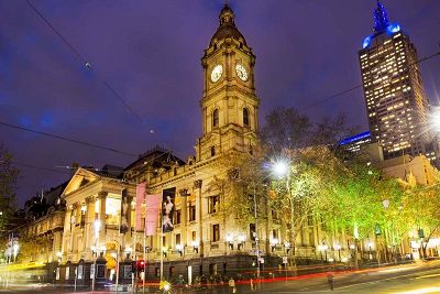 Colour photograph of the Melbourne Town Hall from Swanston Street at night. On the corner of two city streets the building, with its French Renaissance style clock tower prominently positioned, is brightly lit in the evening sky.