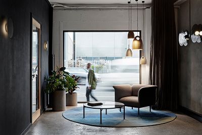 A photo taken from inside the studio looks outside to passersby. The studio is decorated with a circular blue rug, a chair, a table, plants and lights.