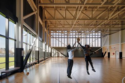 Two students play basketball in the school gymnasium with soft daylight coming in from the glazing that runs along the length of the wall on the left side of the image