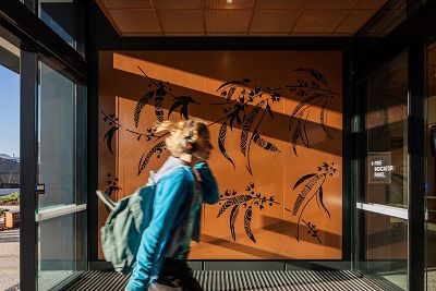 A mid shot of a student entering the foyer of the school with First Nations People artwork featuring along the wall of the entrance. The wall is orange and the student is wearing a light blue top and light green backpack