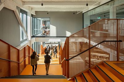 students walking down orange stairs inside the building with views to groups of students in class on the upper level