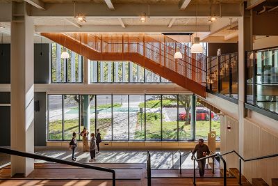 view from the top of the atrium with timber seating and stairs and soft sunlight entering the full height windows of the ground and first floor. The orange stairs stretch across the top half of the image.