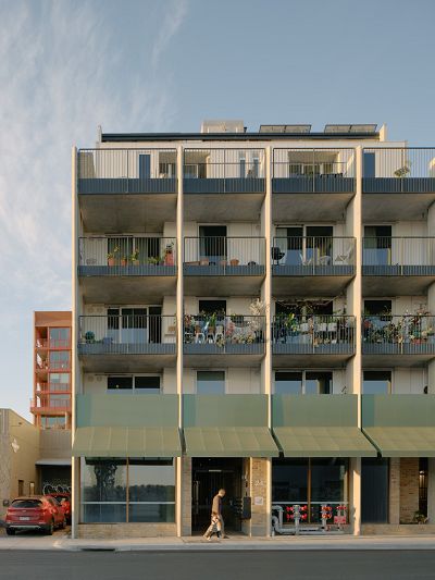 A street view of five storeys of apartments with pea-green awnings protruding over street level and balconies with metal balustrades above that all busy with pot plants and outdoor furniture, a woman with long dark hair and sneakers walks down a concrete open stairway with semicircular transfer decks encircled by pea-green metal balustrades, a minimalist white-painted bathroom with a shower with sparkling brass tapware and a pale green terrazzo tiles on the floor.