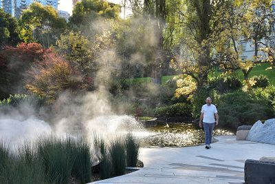 A man is strolling along the stone path which weaves through the memorial. To the left of the man is a fountain that creates a mist cloud that floats above the reeds in the summer light. Behind the man is a garden bed filled with a diverse selection of plants.