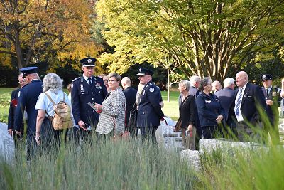 A group of people, some from the Victorian Armed Forces gather together at the Memorial. Surrounding the people is a curved stone wall with name plaques of the people who died in the line of duty. A bank of green reed beds is in the foreground. In the background are trees in autumn colours.