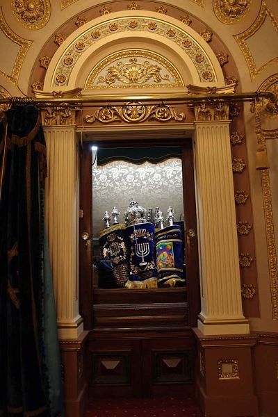 Open ark cabinet holding scrolls covered with decorative velvet covers and silver 'crowns' over winding polls.