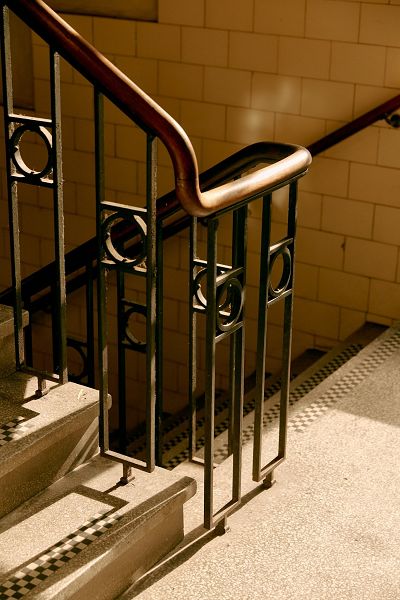 Bottom corner of a stairs case and bannister. Sunlight shines from the right casting shadows of the bannister across the floor and stairs.
