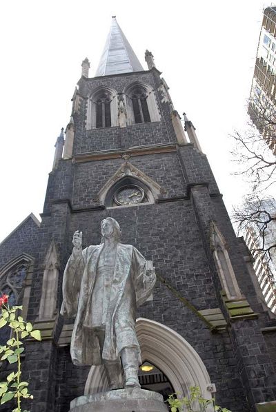 Exterior photograph of a neo-Gothic bluestone church behind a stone statue of a man in robes.