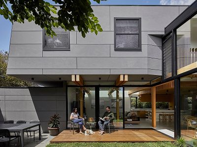 A two-storey home addition clad in grey concrete panels, showing a view to the living room beyond with home-owners relaxing on the deck with their cute white dog