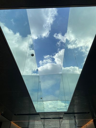 looking up at the skylight of the museum. The skylight is large and surrounded by mirrors, all reflecting blue sky with fluffy white clouds.