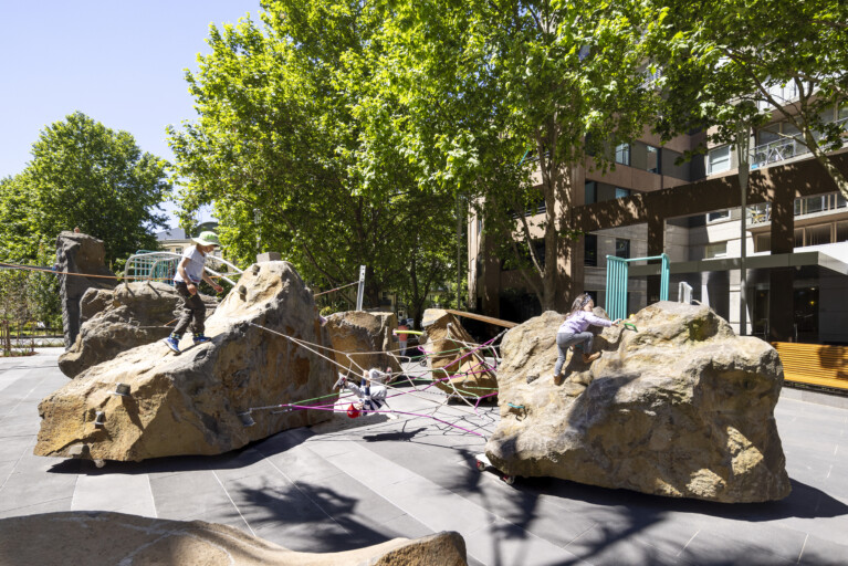 Children climbing a playground which consists of large rocks with various climbing accessories attached such as rope and hand grips. A large green tree shades the playground.